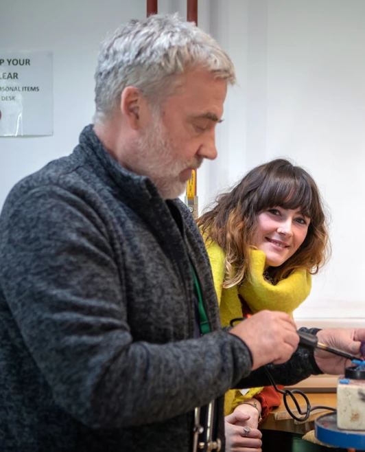 A male instructor with grey hair and a lanyard guiding a female student in a yellow sweater through a jewellery-making process in a workshop setting. A male instructor with grey hair and a lanyard guiding a female student in a yellow sweater through a jewellery-making process in a workshop setting.